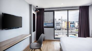 Una habitación de hotel moderna con una cama grande, un televisor de pantalla plana montado en la pared, un escritorio de madera, una silla gris y un amplio ventanal con vistas a la ciudad.