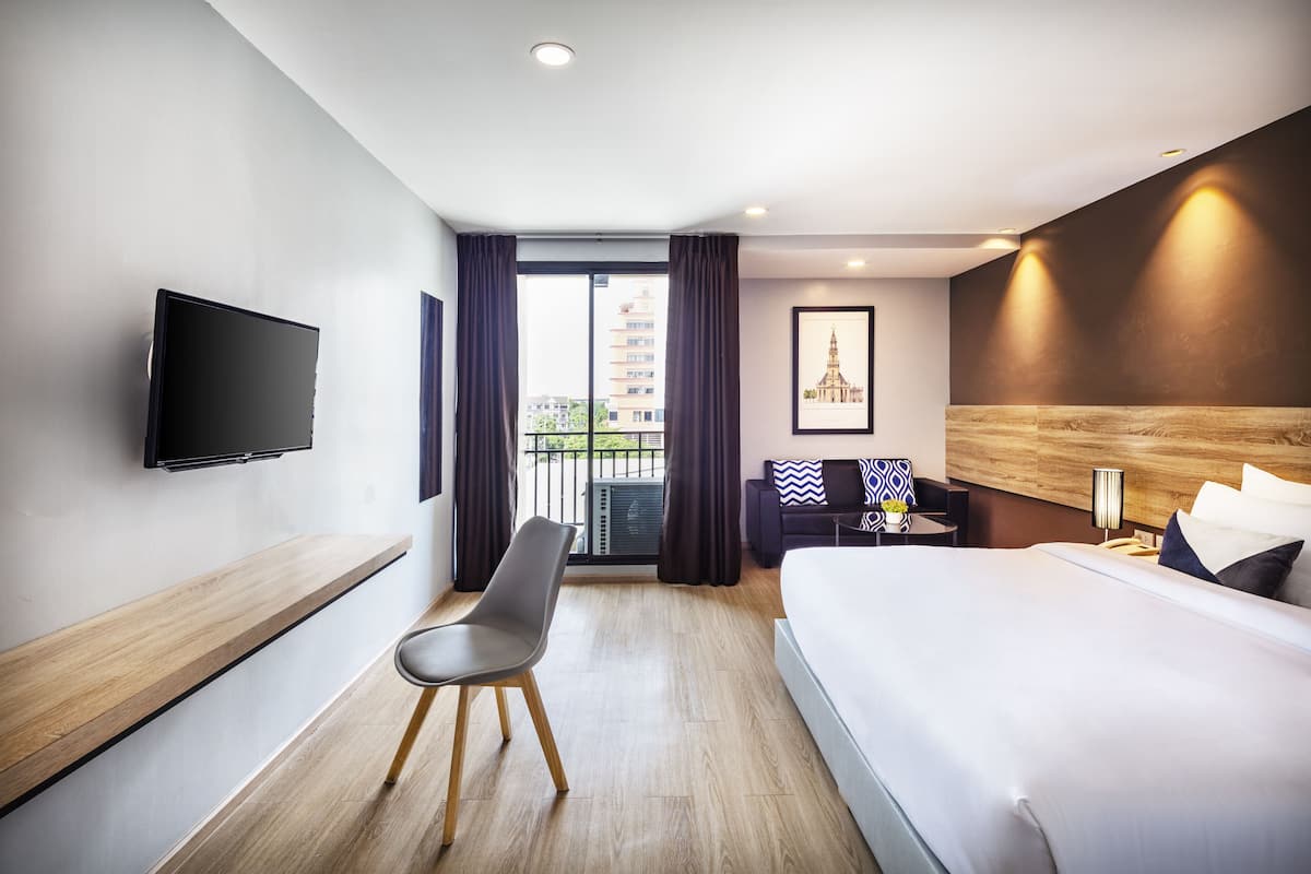 A modern hotel room with a bed, a desk with a chair, a flat-screen TV, a framed picture, and a balcony with curtains.