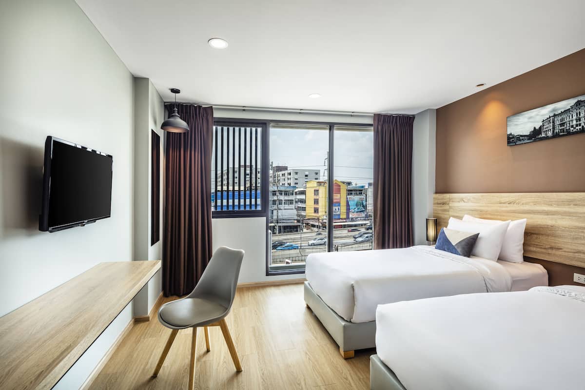 A hotel room with two beds, a desk, a chair, a TV, and a large window with a city view.
