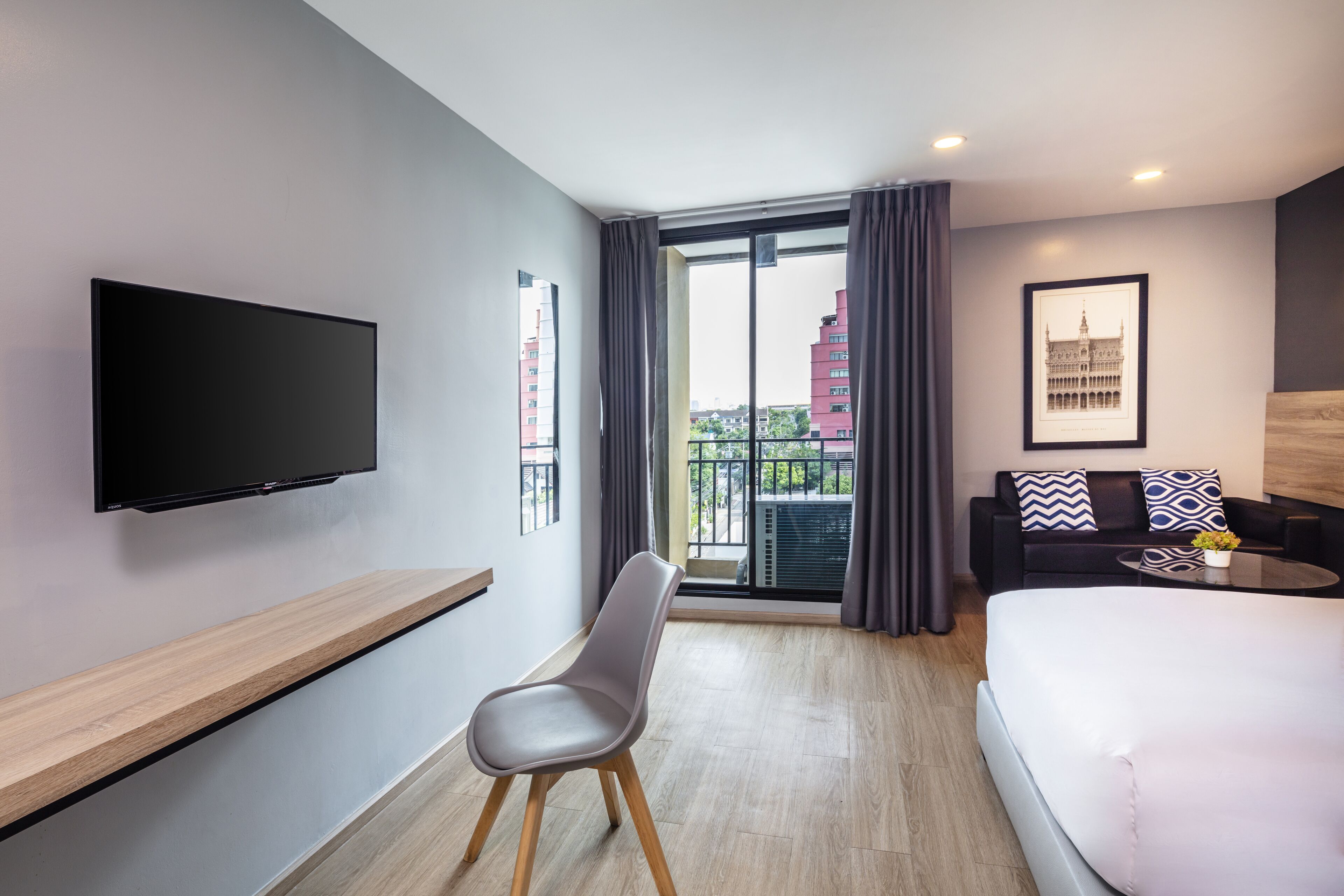 Una habitación de hotel moderna con una cama grande, un televisor de pantalla plana montado en la pared, una zona de estar con un sofá y una silla, y un balcón con vistas a la ciudad.