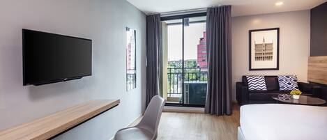 Una habitación de hotel moderna con una cama grande, un televisor de pantalla plana montado en la pared, una zona de estar con un sofá y una silla, y un balcón con vistas a la ciudad.