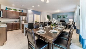 A modern kitchen with dark wooden cabinets, a stainless steel refrigerator, and a dining area with a long wooden table set for six. - Spanish Sunset - Private Pool/Spa & 24/7 Support (Davenport)