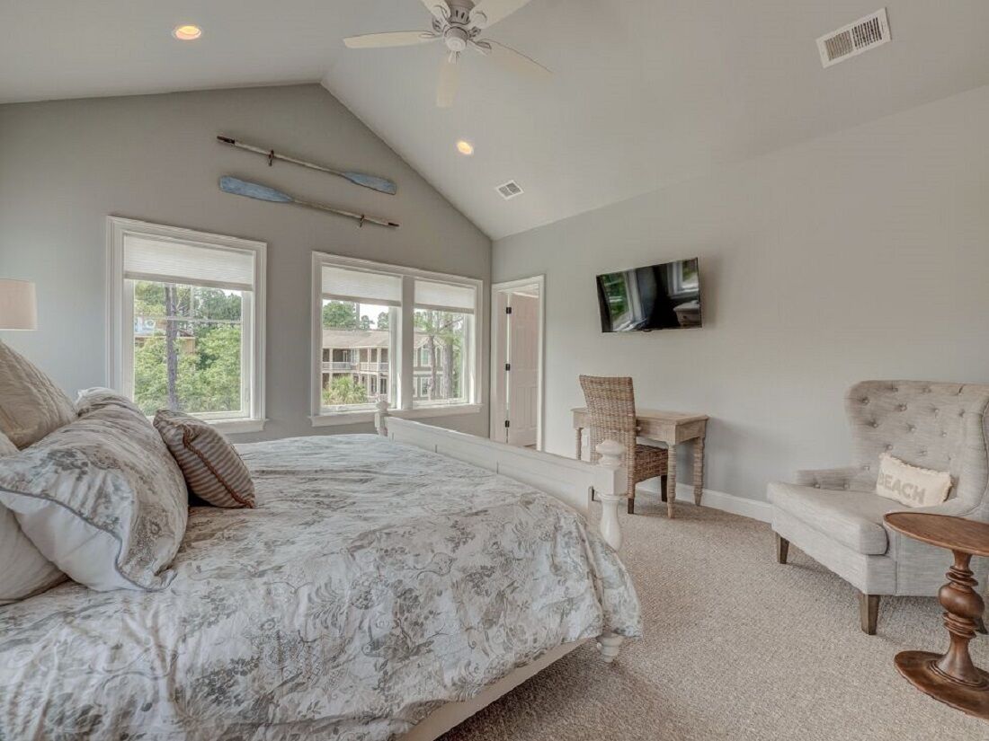 A bedroom with a large bed, a ceiling fan, a TV mounted on the wall, and a view of the outdoors through the windows.