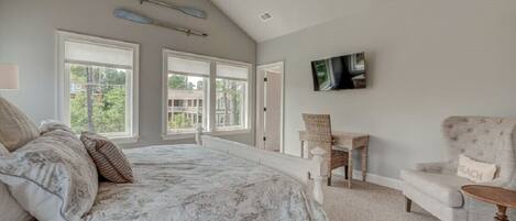 A bedroom with a large bed, a ceiling fan, a TV mounted on the wall, and a view of the outdoors through the windows.