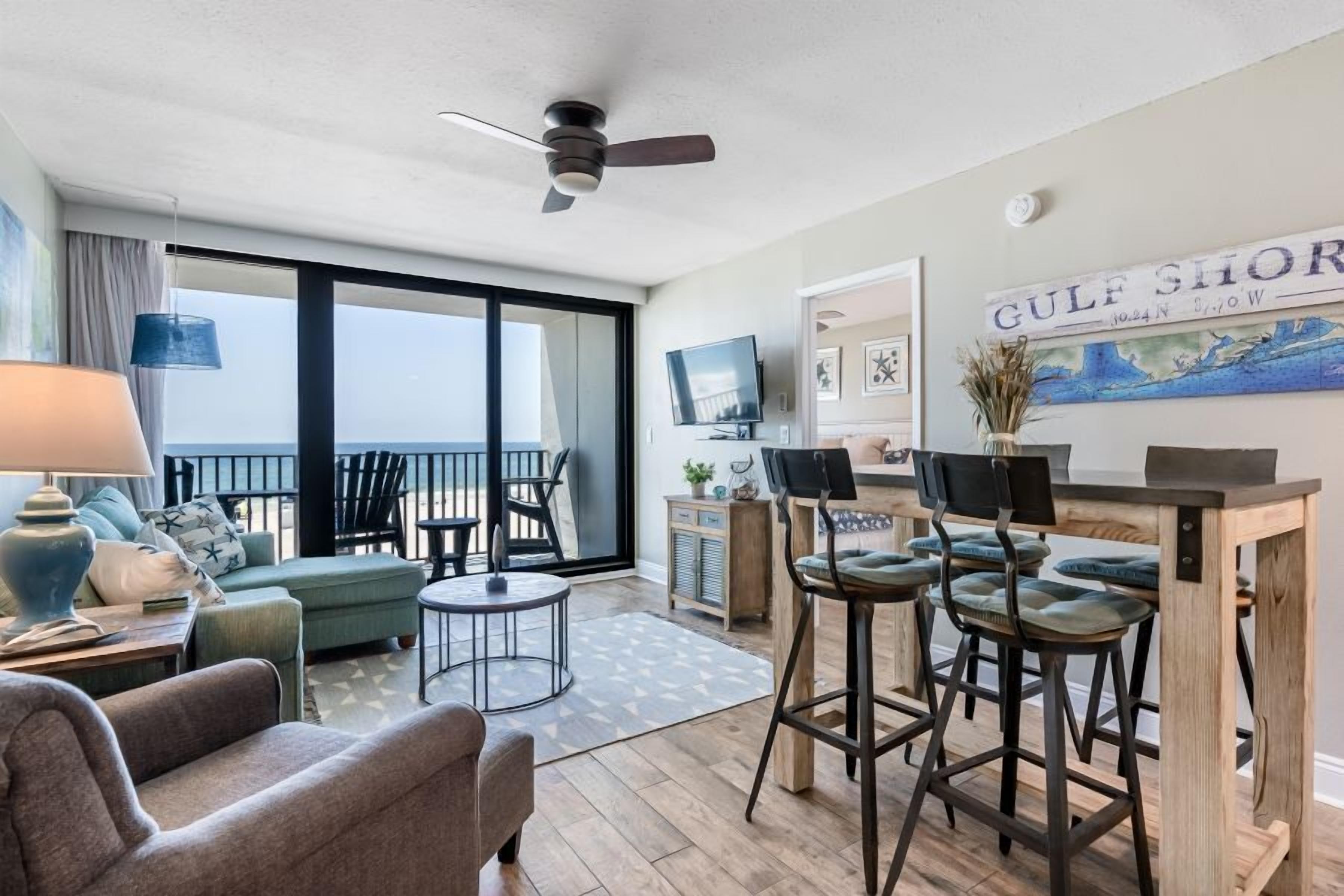 A modern living room with a sofa, chairs, a coffee table, and a dining area with bar stools. There is a large window with a view of the ocean, a ceiling fan, and a television.