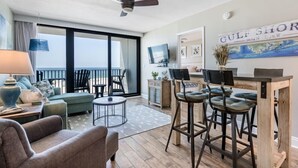 Una sala de estar moderna con un sofá, sillas, una mesa de centro y una zona de comedor con taburetes de bar. Se aprecia una ventana grande con vistas al océano, un ventilador de techo y un televisor.