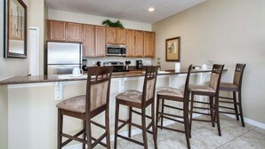 A kitchen with wooden cabinets, a stainless steel refrigerator, a microwave, and a coffee maker. There are four wooden bar stools. - Disney Escape | Private Pool | Resort Amenities | Sleeps 10 (Kissimmee)