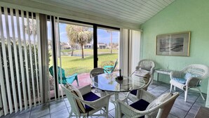 Un patio techado con una mesa de vidrio rodeada de sillas de mimbre, un ventilador de techo y vistas a un césped verde y palmeras.