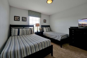 A room with two beds, a window with blinds, a lamp, and a television on a stand.