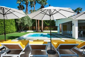 Et poolområde med liggestole og parasoller, en pool samt en bygning med moderne arkitektur.
