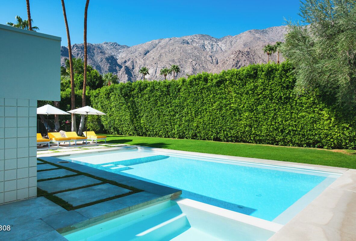 A swimming pool with a view of mountains, surrounded by a hedge and a building with lounge chairs.