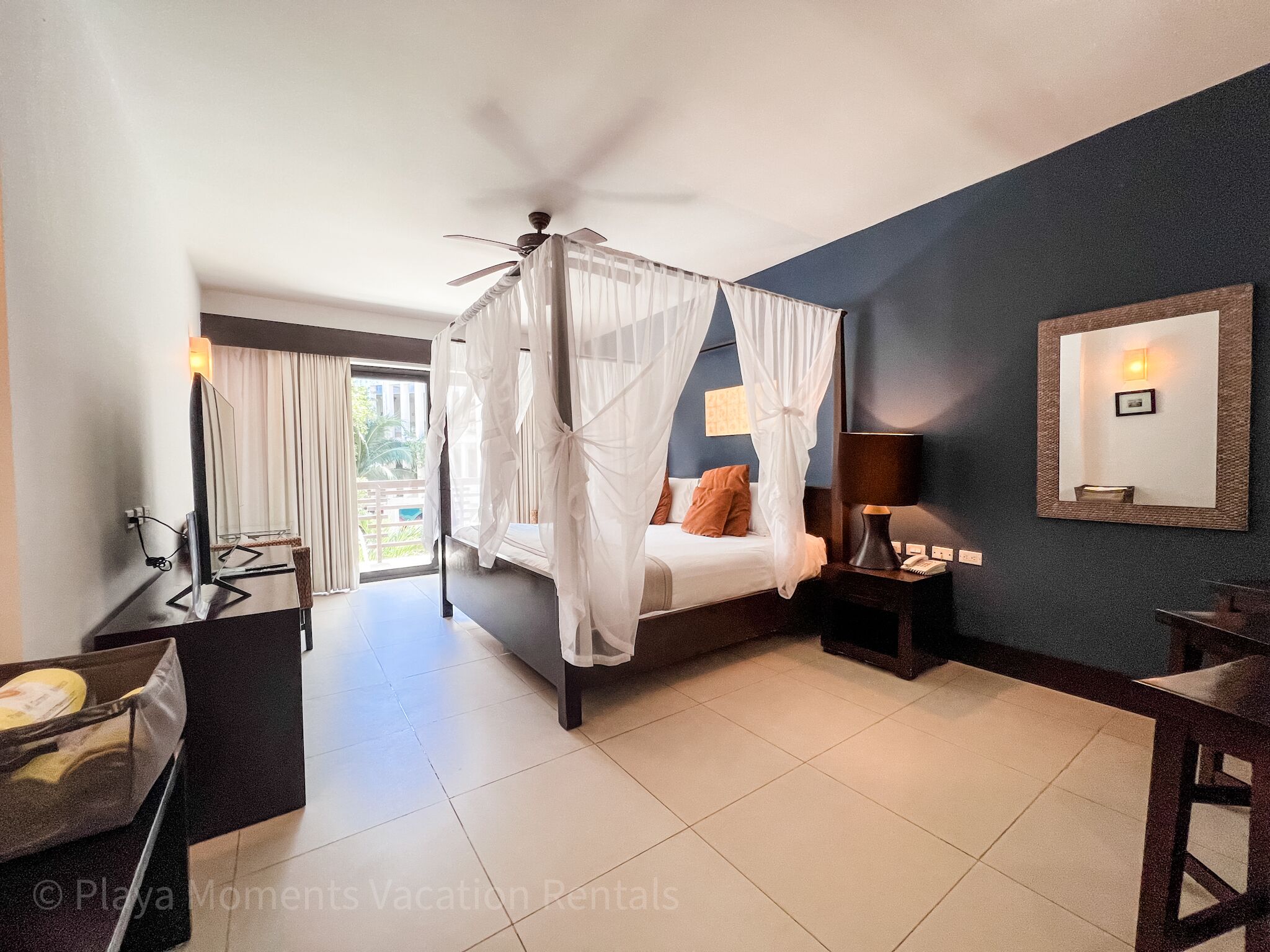 A bedroom with a four-poster bed, a ceiling fan, a bedside table with a lamp, and a large mirror on the wall.