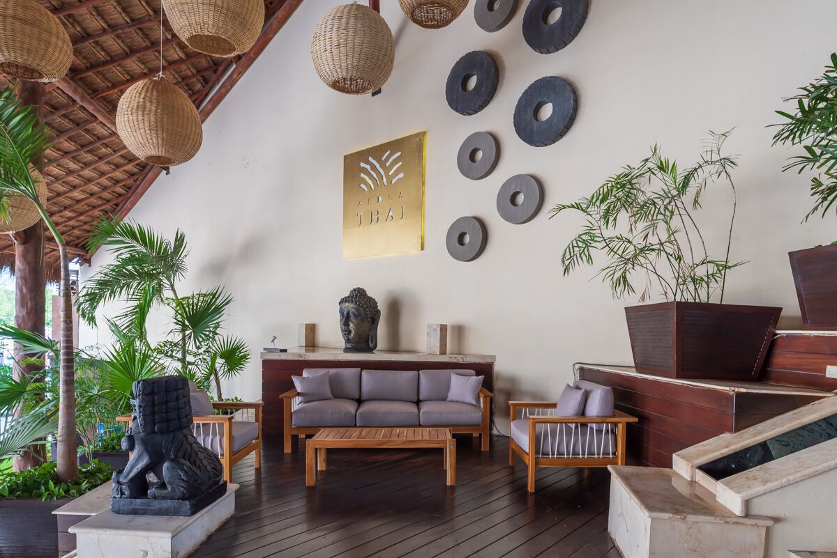 Et moderne loungeområde med en træsofa, et sofabord og en Buddha-statue. Der er vævede lamper, et Buddha-hoved monteret på væggen og en stor plantekasse med bambus.