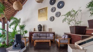 Een moderne zithoek met een houten bank, een salontafel en een Boeddhabeeld. Er zijn geweven lampen, een aan de muur bevestigd Boeddha hoofd en een grote plantenpot met bamboe.