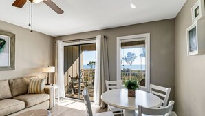 Ein modernes Wohnzimmer mit einer Couch, einem Esstisch mit Stühlen, einem Deckenventilator und Meerblick durch große Fenster.