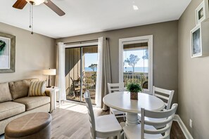 Un salon moderne avec un canapé, une table à manger et des chaises, un ventilateur de plafond et une vue sur la mer grâce à de grandes fenêtres.