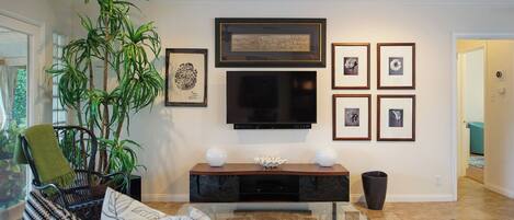 Una sala de estar con un sofá, una mesa de centro, un televisor, cuadros enmarcados y una planta grande.