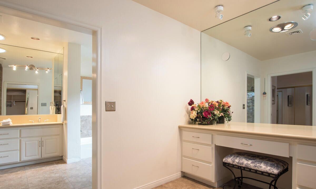 A modern bathroom with a large mirror, a vanity with drawers, a bench, and a floral arrangement.