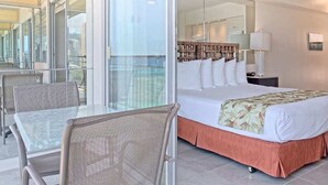 Habitación de hotel con una cama grande, un balcón con paredes de vidrio, una zona de comedor con mesa y sillas, y vistas a la ciudad.