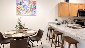 Eine moderne Küche mit Holzschränken, einer Granit Arbeitsplatte und Edelstahlgeräten. Es gibt einen Essbereich mit Tisch und Stühlen, sowie ein farbenfrohes Bild an der Wand.