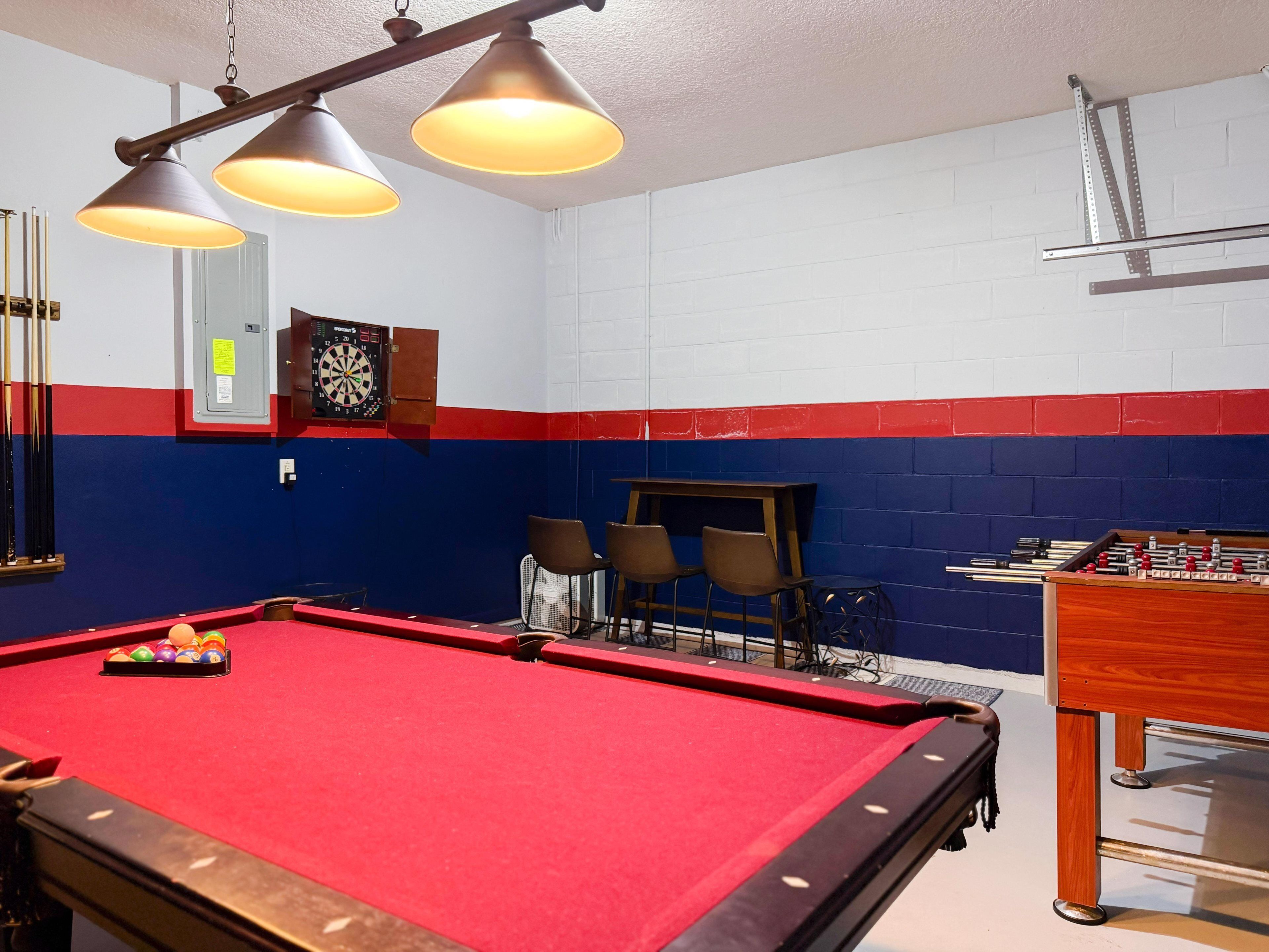 Une table de billard recouverte d’un drap de feutre rouge, trois lampes suspendues et un jeu de fléchettes accroché au mur.
