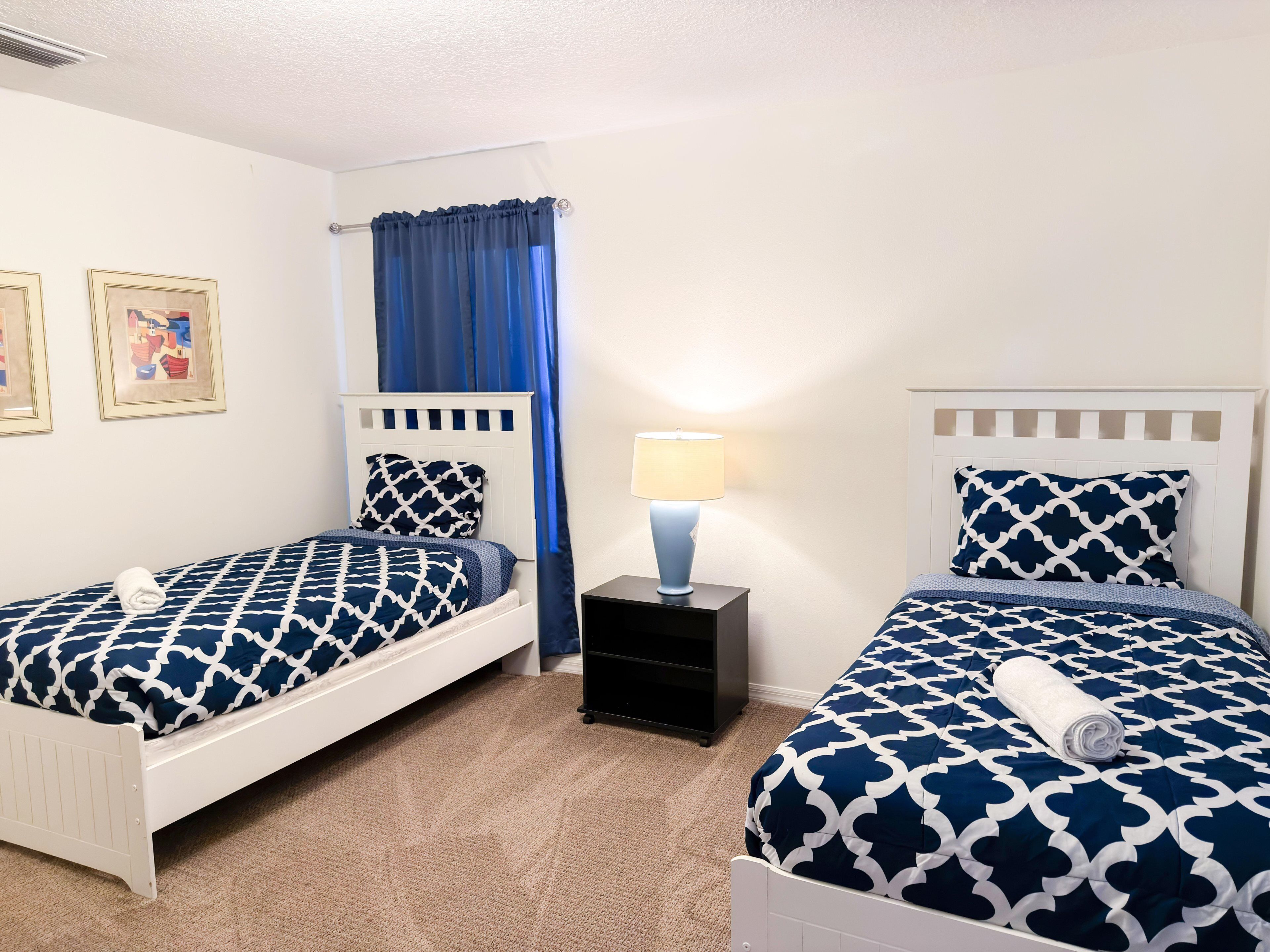 A bedroom with two beds, a nightstand, a lamp, and a window with blue curtains.