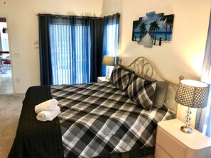 A neatly made bed with a checkered bedspread, a pair of rolled towels, a metal-framed headboard, a bedside lamp, and a wall-mounted artwork of a beach scene. - Disney Area - 4 bedrooms / 2 bathrooms pool home with golf view (Davenport)