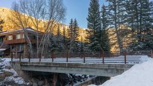 A bridge over a snow-covered stream with a wooden building on the left and tall evergreen trees in the background. - Riverside C03 A Mountain Forest Hideaway - 2 Bedrooms, 2 Bathrooms, Sleeps 6 (Telluride)