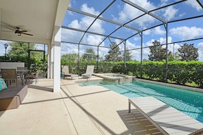 A covered outdoor swimming pool with lounge chairs and a table, surrounded by greenery.