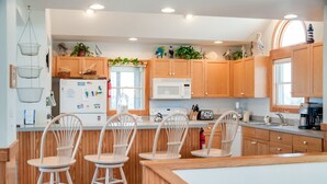 A modern kitchen with wooden cabinets, a white refrigerator, a microwave, and a sink. There are three white chairs and a small island with a sink.