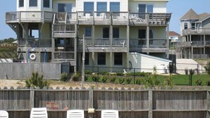 A multi-story beachfront house with balconies, a wooden deck, and a fenced front yard.