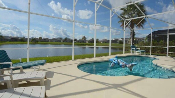 A covered outdoor swimming pool with lounge chairs and a view of a lake and palm trees.
