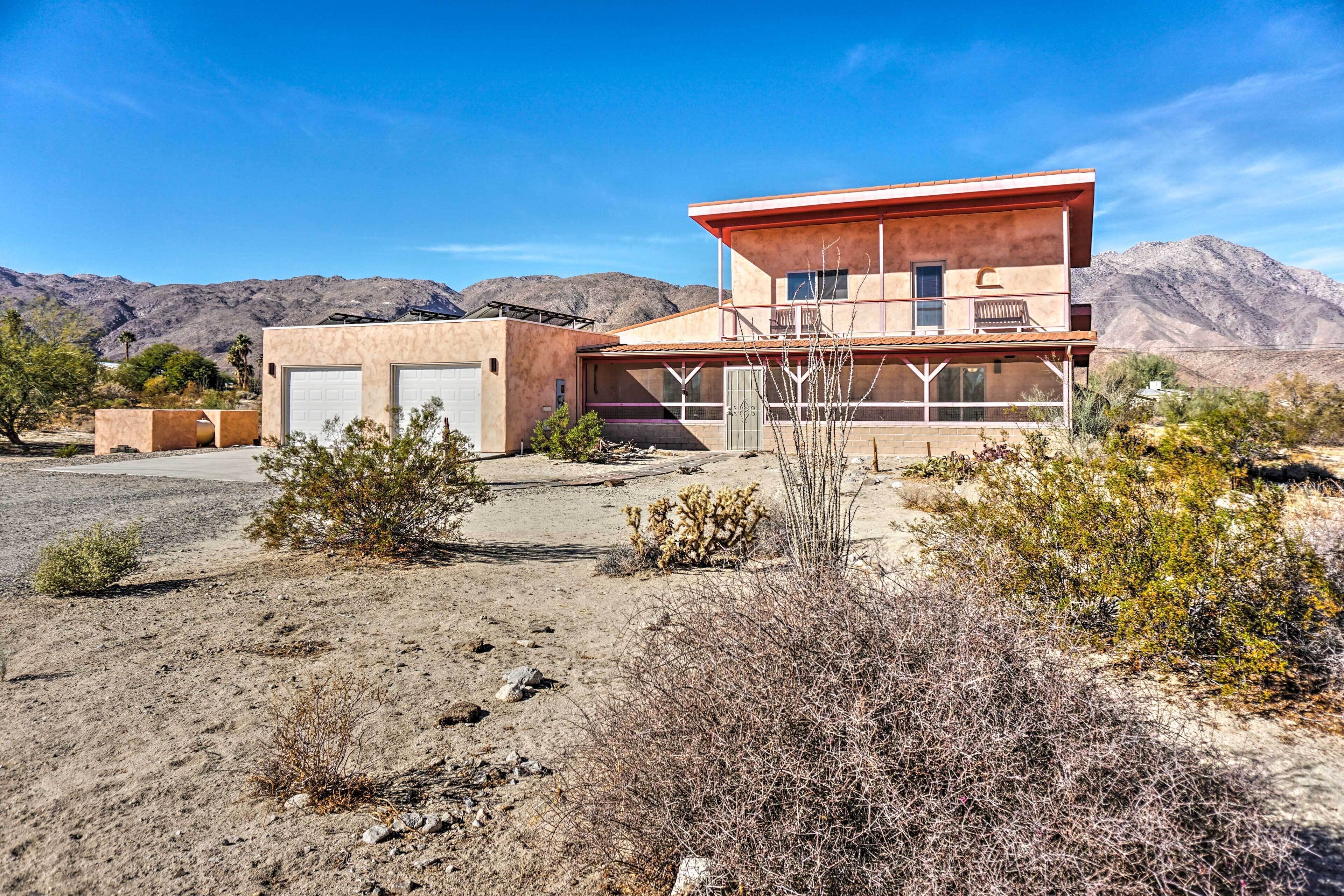 Une maison moderne à deux étages avec un garage, entourée d’une végétation désertique et avec des montagnes en arrière-plan.