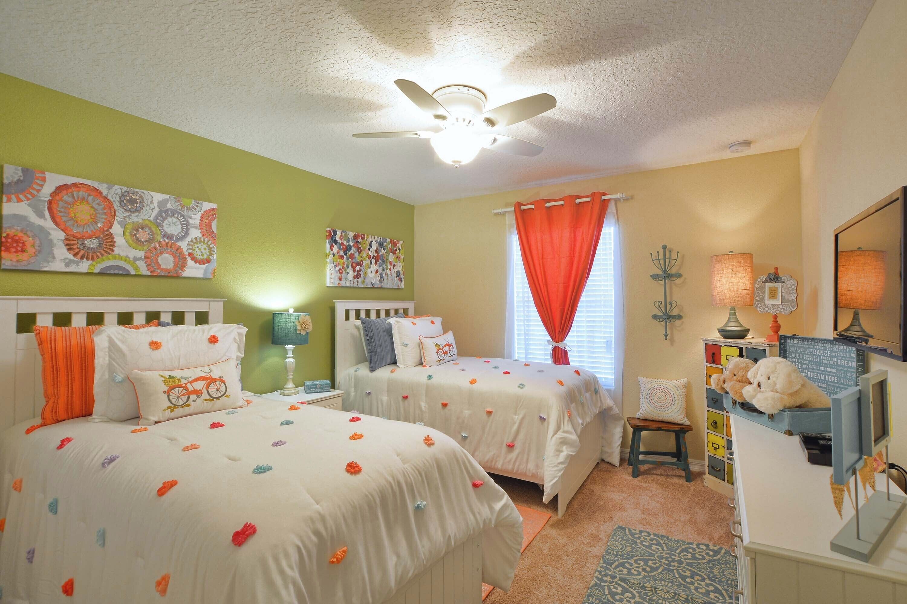 A bedroom with two beds, a ceiling fan, a window with red curtains, and a dresser with a mirror and decorative items.