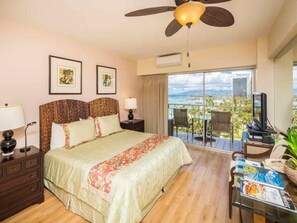 A hotel room with a large bed, a ceiling fan, a balcony with a view of the city, and a desk with a computer and books. - Modern Meets Tropics! Ocean View Suite w/WiFi, Kitchenette – Waikīkī Shore 1005 (HONOLULU)