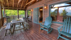 A wooden deck with a picnic table and Adirondack chairs, large windows, and a view of trees.