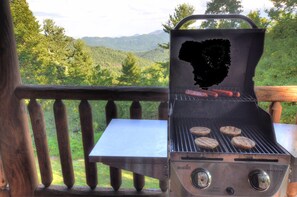 Una parrilla con salchichas y hamburguesas en una terraza de madera con vista a una zona boscosa.