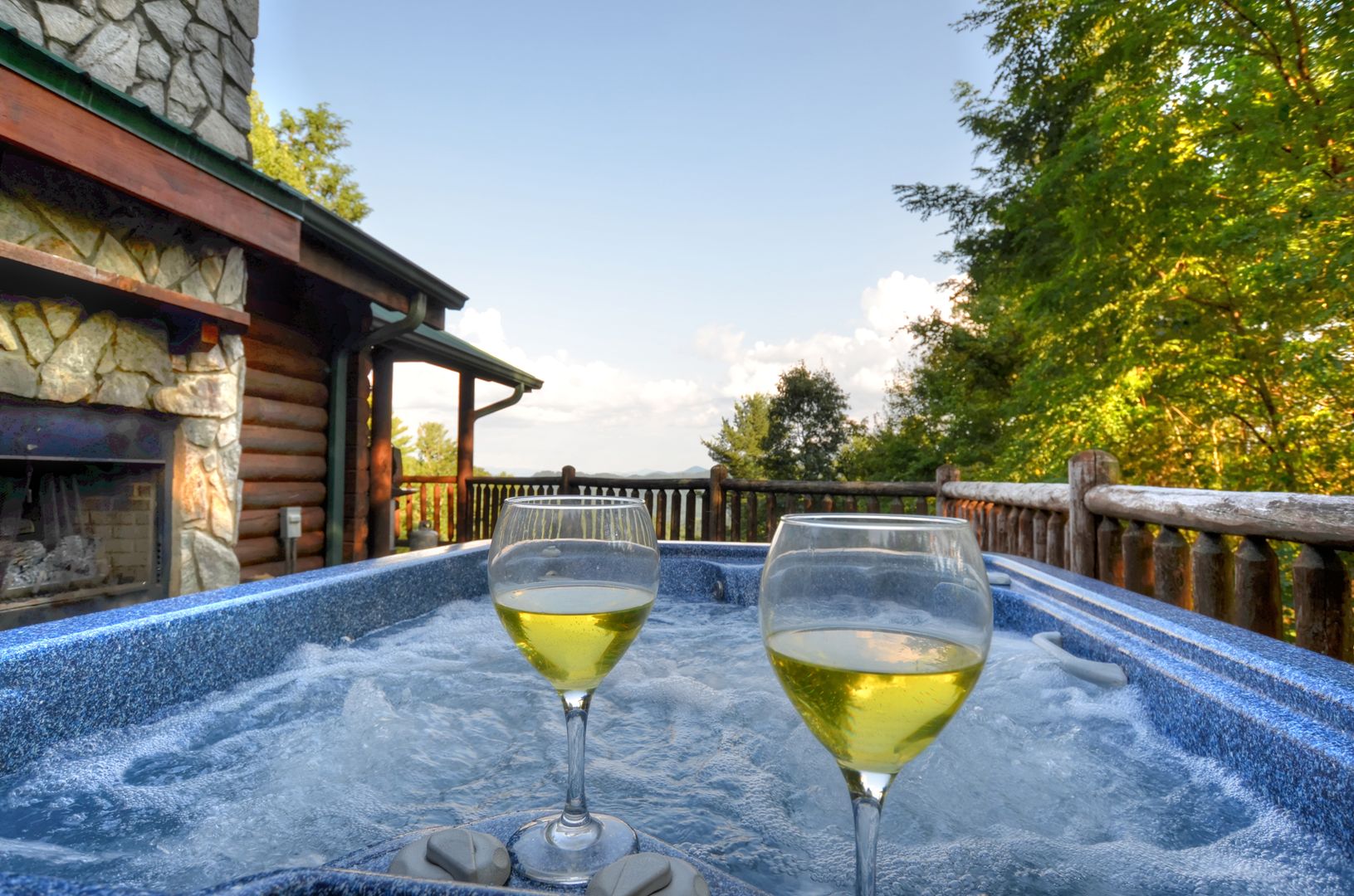 Two glasses of white wine on a hot tub with a stone wall and wooden fence in the background.