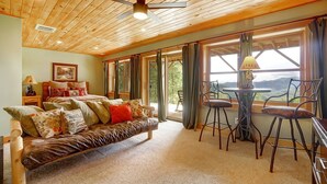 A cozy room with a wooden ceiling, a sofa with patterned cushions, a table with chairs, and large windows offering a view of the outdoors.