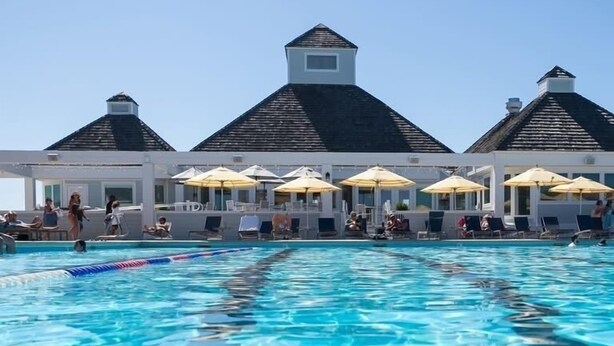 A swimming pool with clear water, lounge chairs, and umbrellas under a building with a peaked roof.