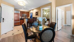 A modern kitchen with stainless steel appliances, a dining area with a glass table and chairs, and a hallway with white doors.