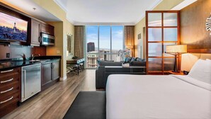 A modern hotel room with a large bed, a desk, and a kitchenette. The room has a city view.