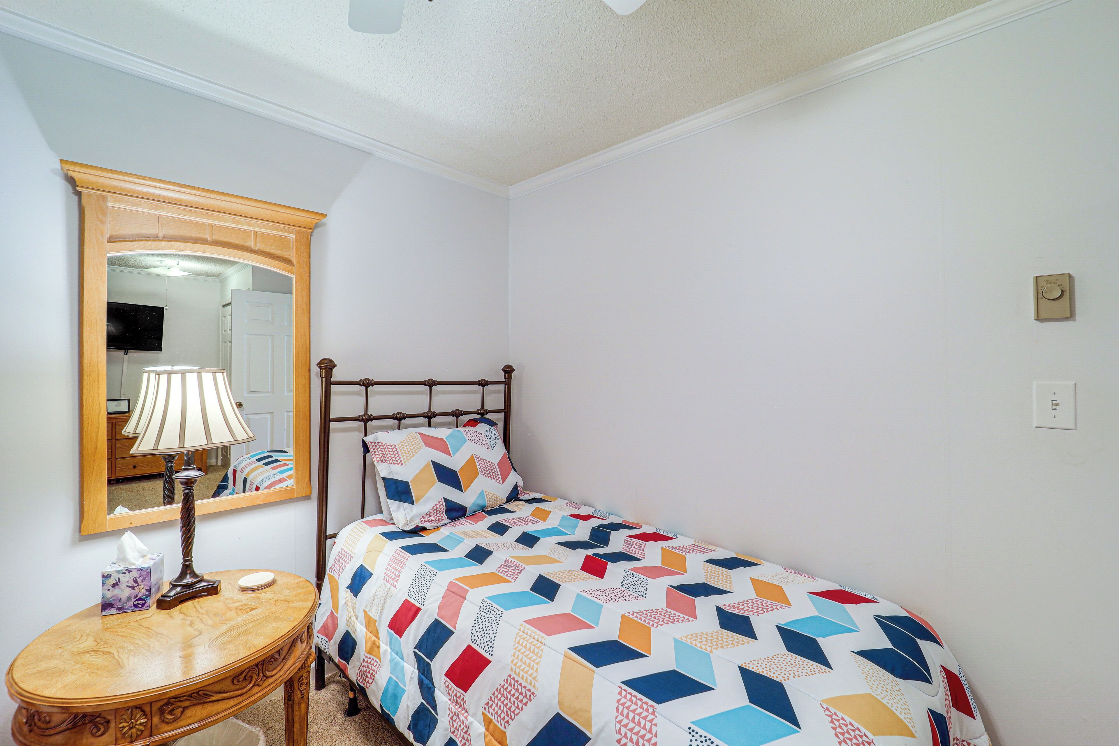 A small, neatly arranged bedroom with a single bed, a bedside table with a lamp, a mirror, and a wall-mounted light switch.