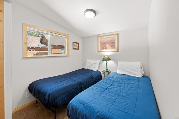 Ein Schlafzimmer mit zwei Einzelbetten, einem Fenster mit Blick auf ein Haus, einem gerahmten Bild an der Wand und einer Lampe auf einem Nachttisch.