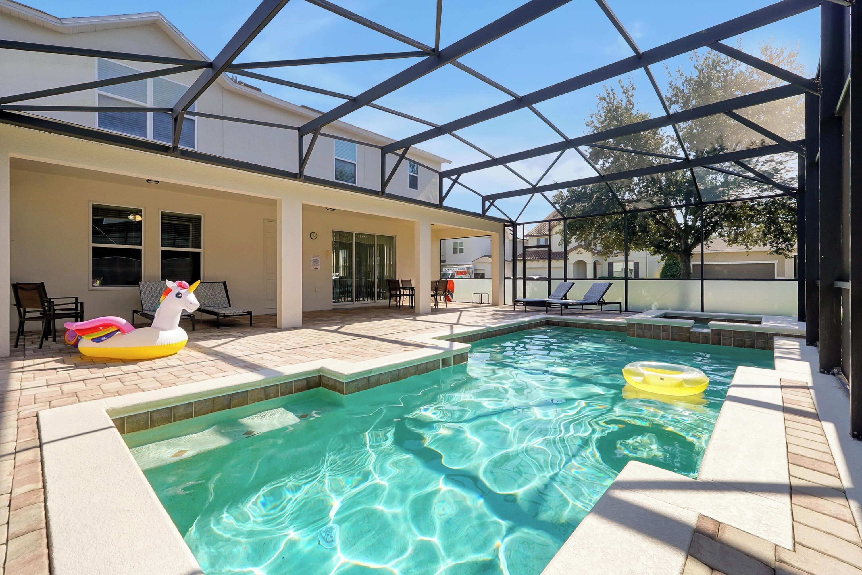 A covered outdoor swimming pool with inflatable toys, a patio area with chairs, and a view of neighboring houses.