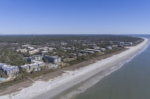 An aerial view of a coastal area with a long beach, several multi-story buildings, and a dense forested area.