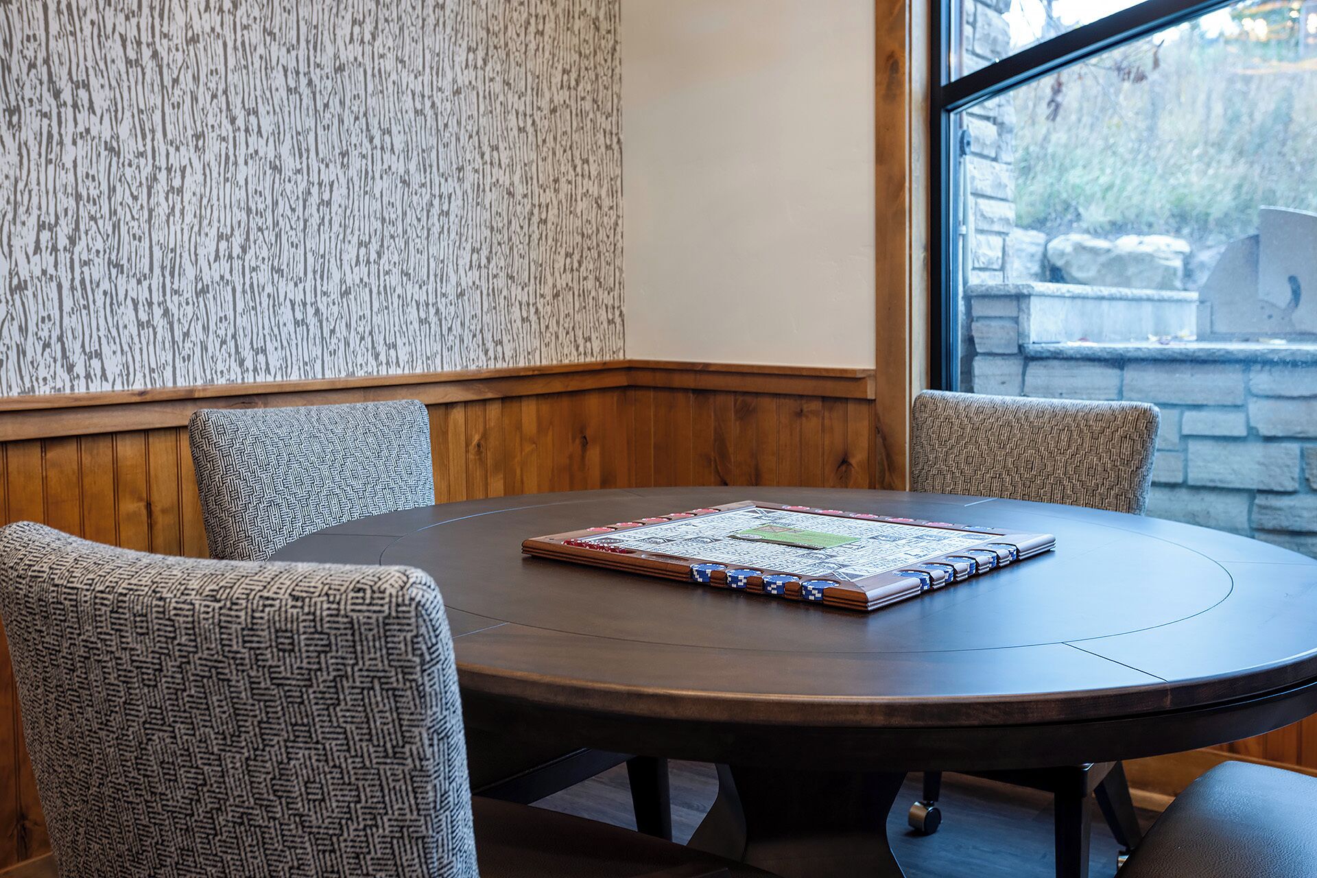 A round table with a board game, four patterned chairs, a wooden wall with a vertical pattern, and a stone wall visible through a window.