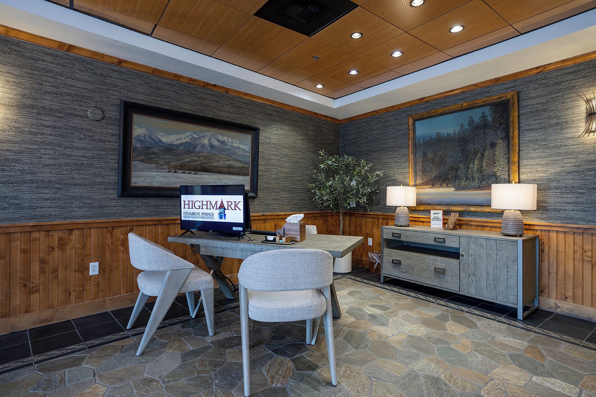 A hotel lobby with a reception desk, two chairs, a television screen, and a large painting on the wall.