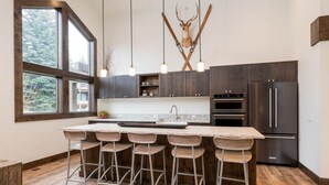 A modern kitchen with a large island, bar stools, dark wood cabinets, stainless steel appliances, and a deer head mounted on the wall.