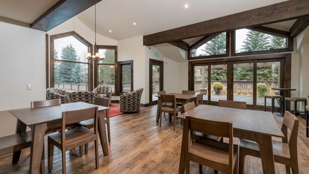 A spacious dining area with wooden tables and chairs, large windows offering views of trees, and a cozy seating area with patterned cushions.